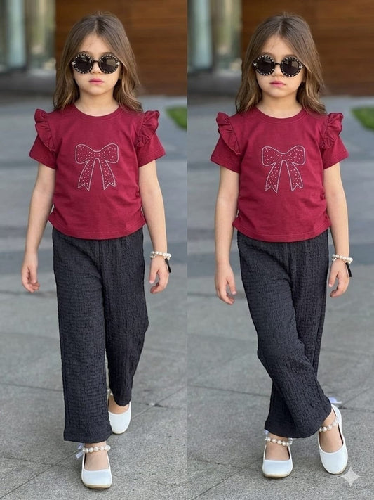 A girl wearing a The Nile Luxuries burgundy ruffle-sleeve top with a sparkling bow and black textured trousers.