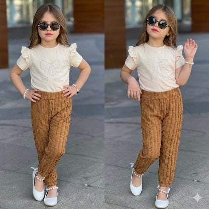 The Nile Luxuries girls' outfit with a cream ruffle-sleeve bow t-shirt and brown textured trousers, worn by a child.