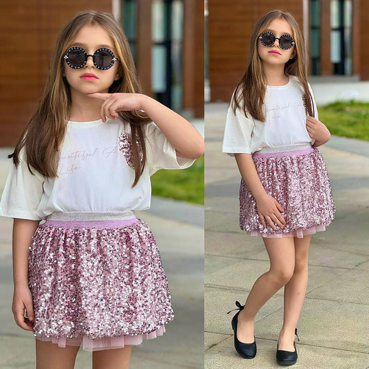 The Nile Luxuries girls' outfit with a white graphic t-shirt and pink sequin tulle skirt, worn by a young girl.