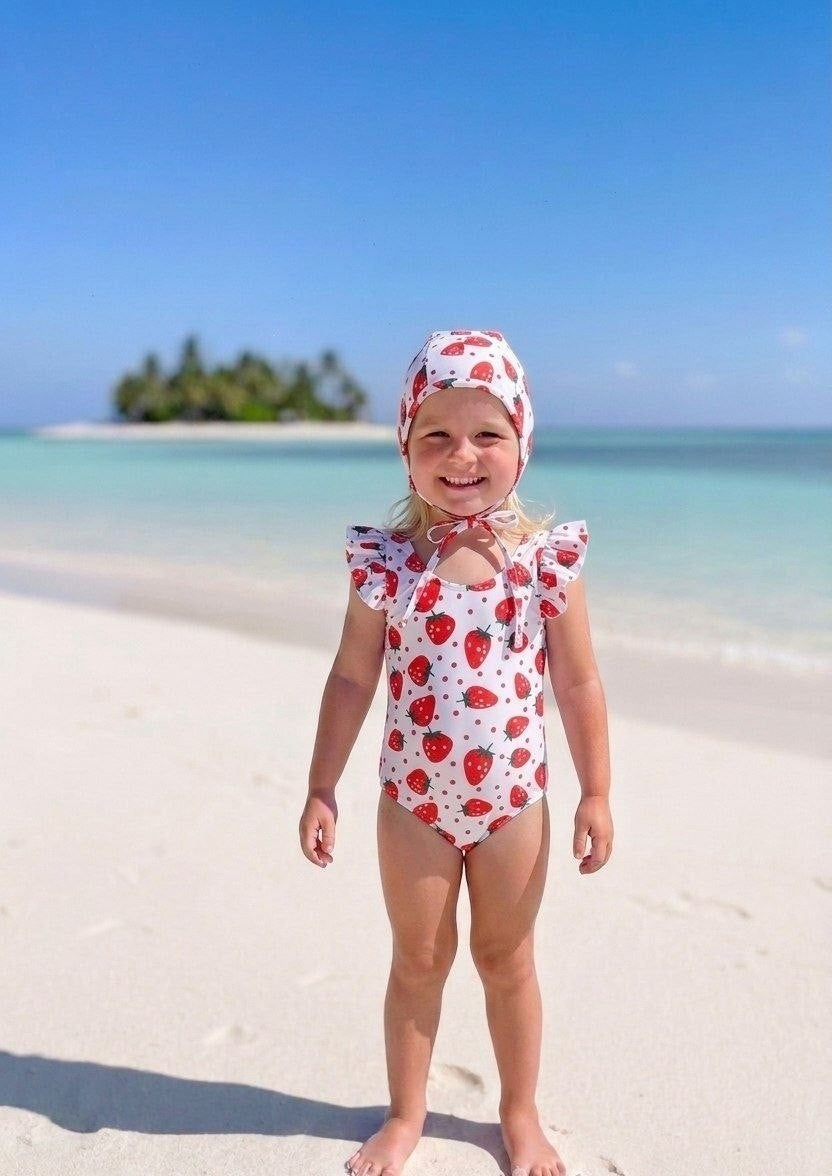 The Nile Luxuries girls' white swimsuit with red strawberry print and ruffle sleeves, matching bonnet. Beach setting.