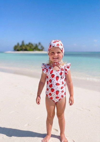The Nile Luxuries girls' white swimsuit with red strawberry print and ruffle sleeves, matching bonnet. Beach setting.