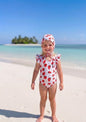 The Nile Luxuries girls' white swimsuit with red strawberry print and ruffle sleeves, matching bonnet. Beach setting.