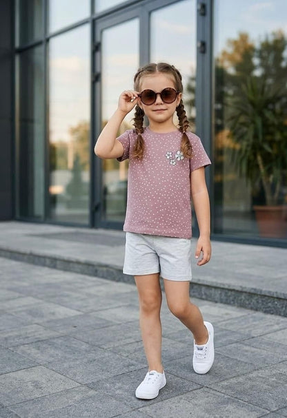A young girl wearing a The Nile Luxuries mauve polka dot t-shirt with floral appliqué and grey shorts outdoors.