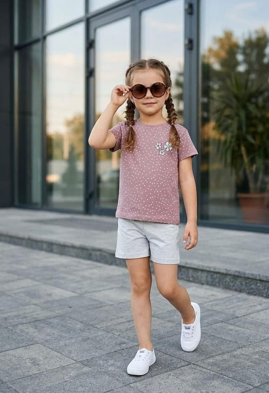 A young girl wearing a The Nile Luxuries mauve polka dot t-shirt with floral appliqué and grey shorts outdoors.