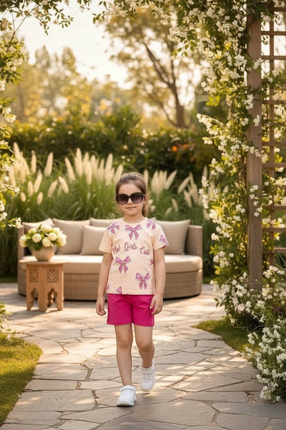 The Nile Luxuries girls' summer set with a peach bow-print t-shirt and pink shorts, worn by a girl in a garden.