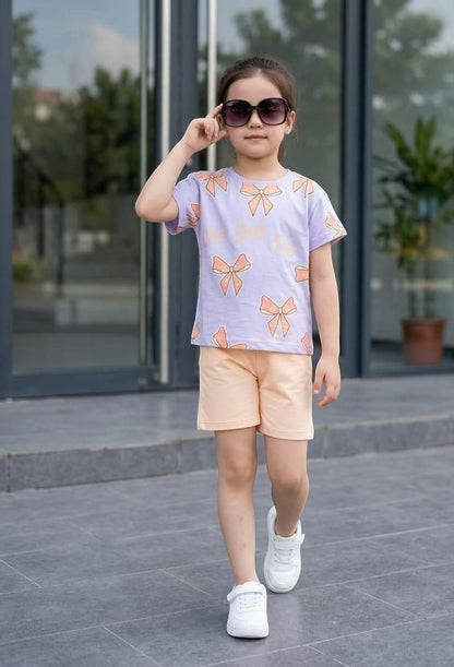 Young girl wearing The Nile Luxuries lavender bow-print t-shirt and peach shorts with white sneakers and sunglasses.