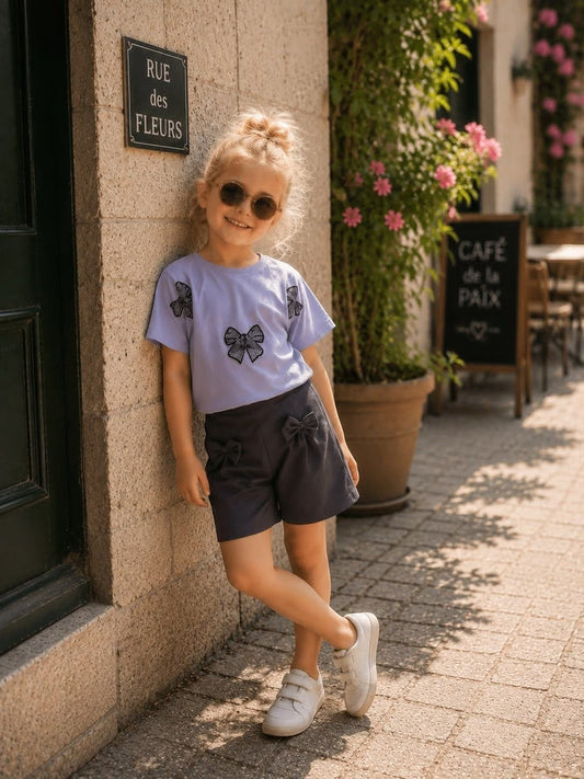 The Nile Luxuries girls summer outfit with a light blue bow-print t-shirt and dark grey shorts worn by a young girl.