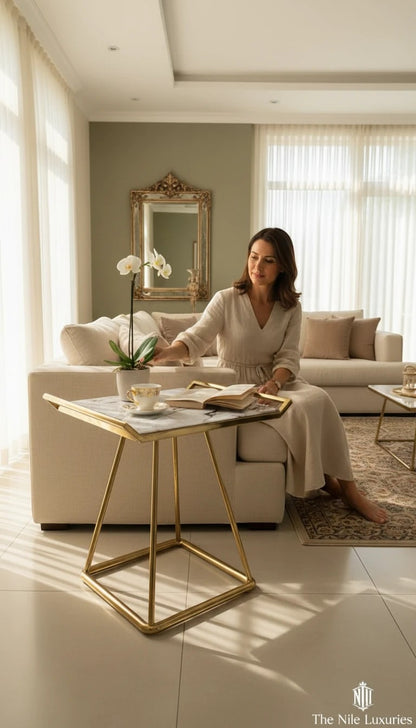 The Nile Luxuries modern accent table with white marble-look top and gold geometric base. A woman sits beside it.