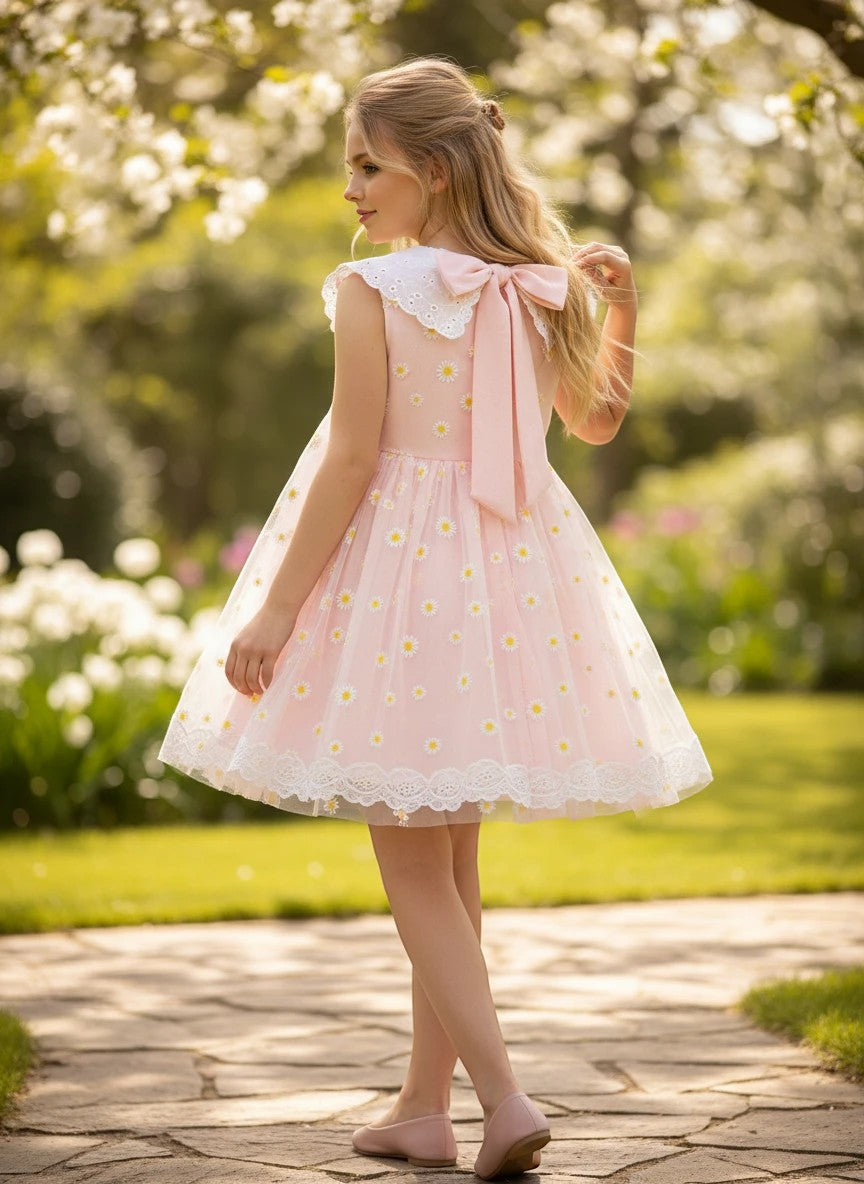 The Nile Luxuries pink tulle dress with daisy embroidery, Peter Pan collar, and oversized bow, worn by a girl outdoors.