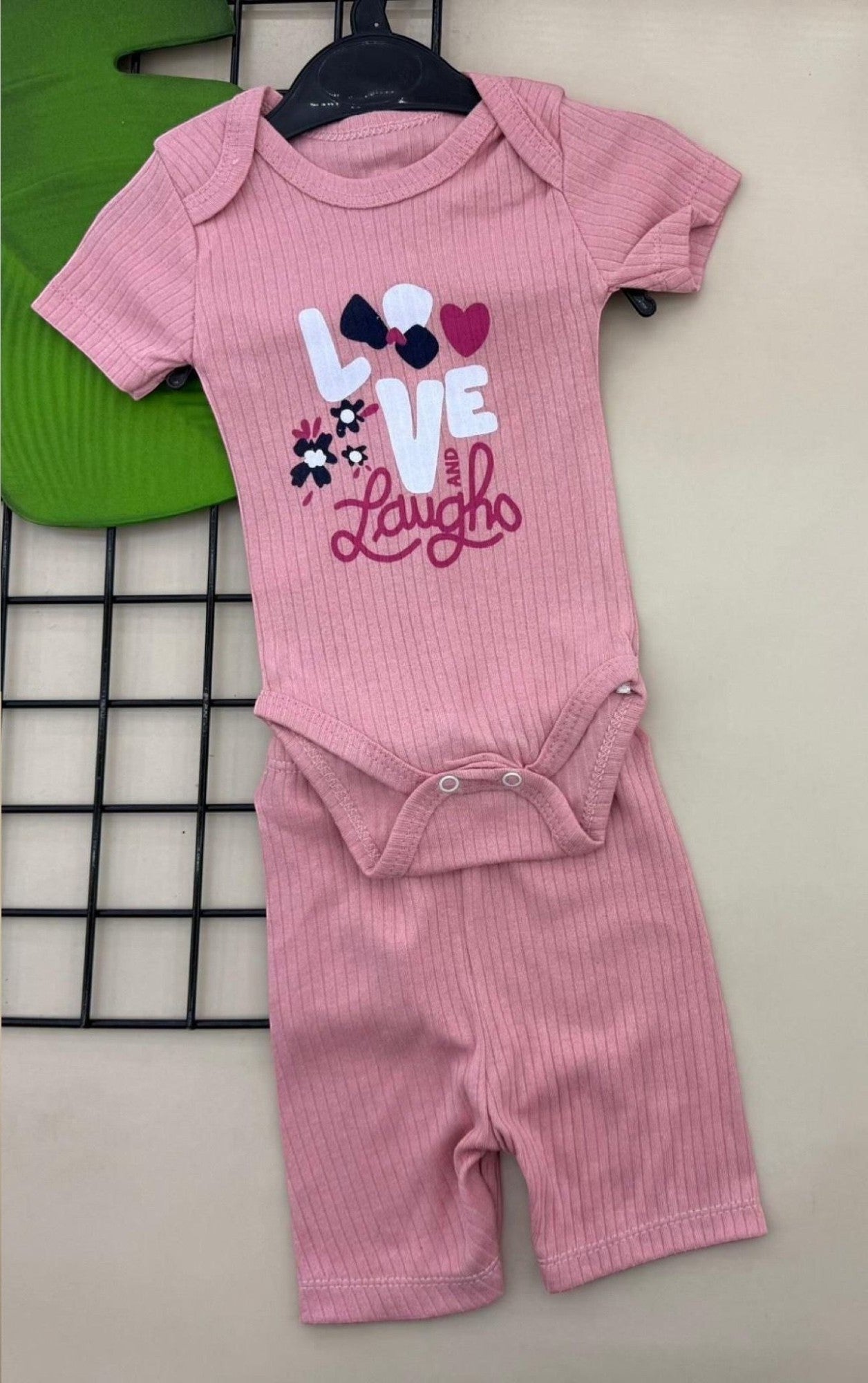 The Nile Luxuries pink ribbed knit baby bodysuit and shorts set with "LOVE and Laugh" graphic and bow detail.
