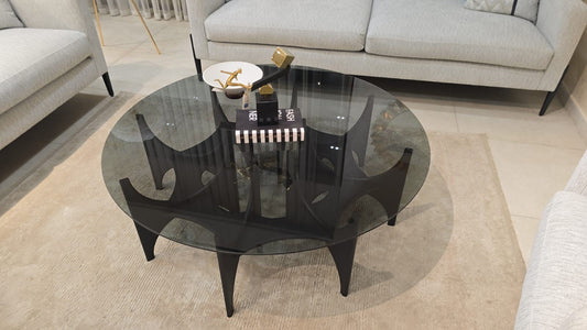 The Nile Luxuries sculptural coffee table with a round smoked glass top and a dark matte base with intricate legs.