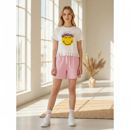 The Nile Luxuries white t-shirt with fringed hem and smiley face graphic, paired with pink shorts. Model wears white sneakers and pink-striped socks.