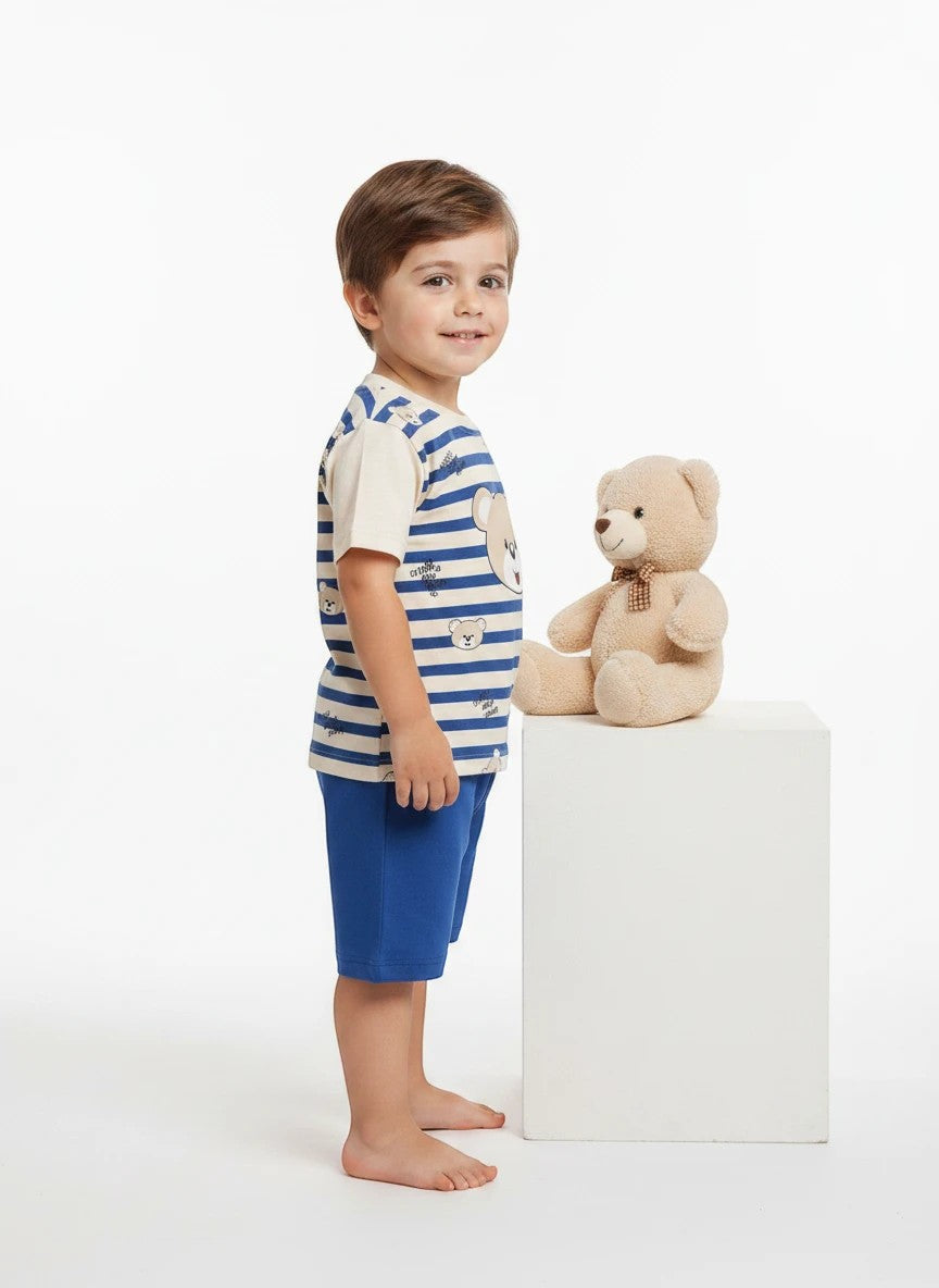 The Nile Luxuries toddler boy wearing a striped bear print t-shirt and blue shorts standing next to a teddy bear.