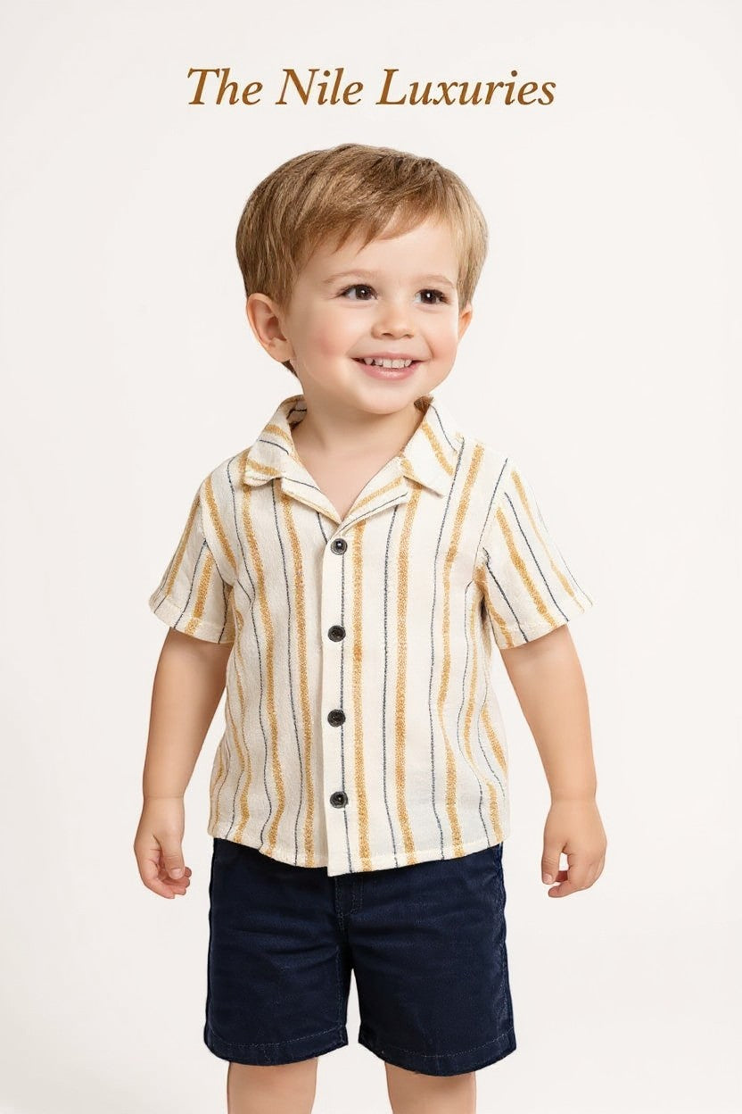 Toddler boy wearing The Nile Luxuries striped button-down shirt and navy shorts, smiling against a white background.