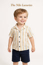 Toddler boy wearing The Nile Luxuries striped button-down shirt and navy shorts, smiling against a white background.