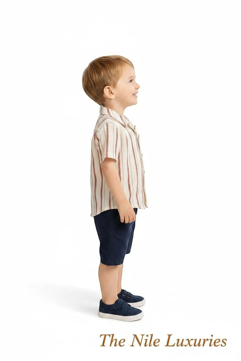 Toddler boy wearing The Nile Luxuries striped button-down shirt and navy shorts, standing against a white background.