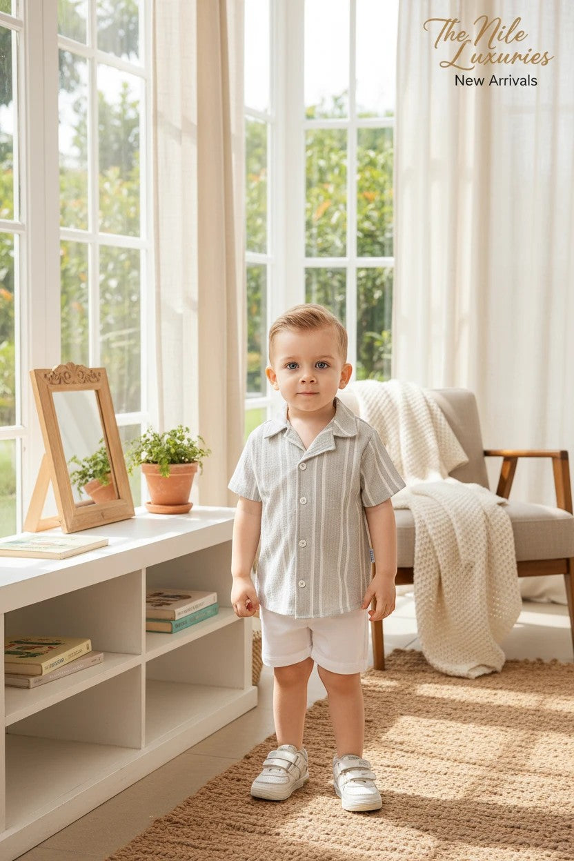 Toddler boy wearing a The Nile Luxuries sage and white striped button-down shirt and white shorts in a bright room.