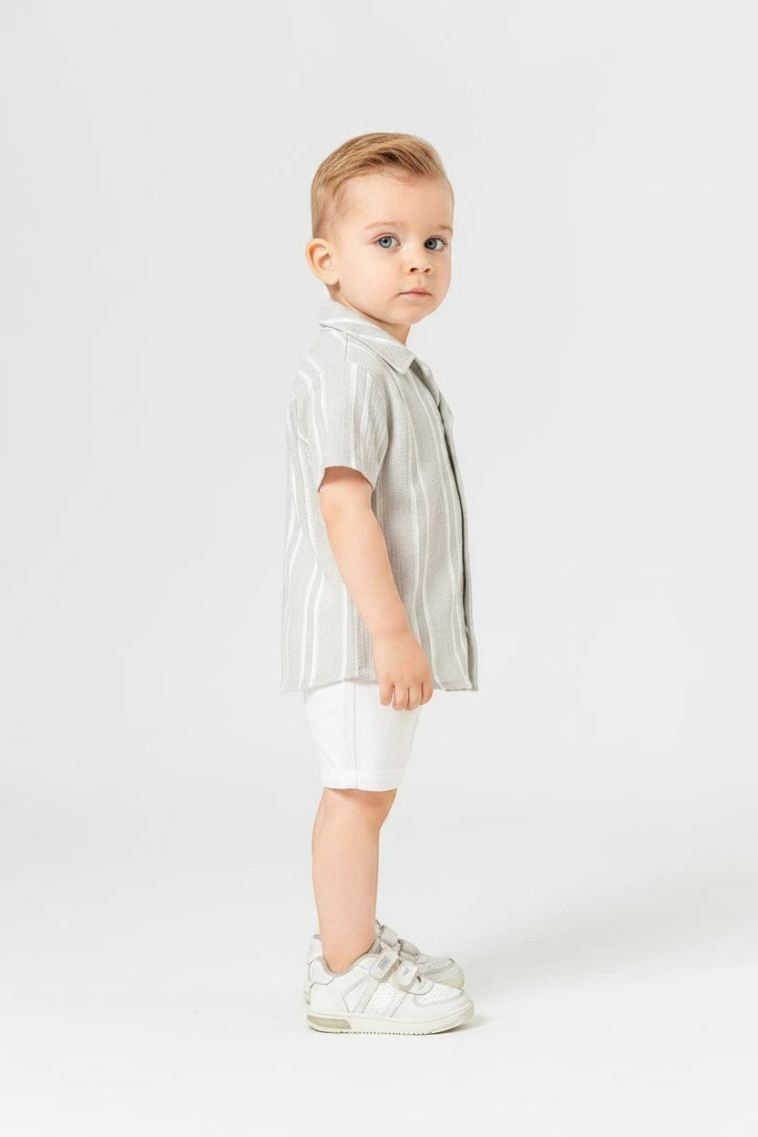 Toddler boy wearing The Nile Luxuries sage striped button-down shirt and white shorts on a white background.