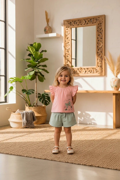 Toddler girl wearing The Nile Luxuries pink flutter sleeve top with butterfly print and sage green bow shorts.