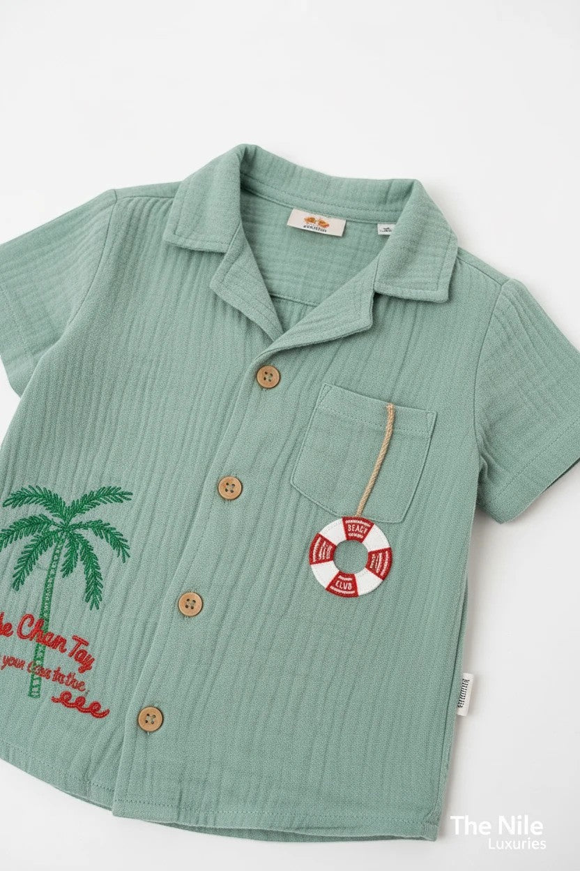 The Nile Luxuries sage green muslin toddler shirt with palm tree embroidery and nautical lifebuoy charm detail.