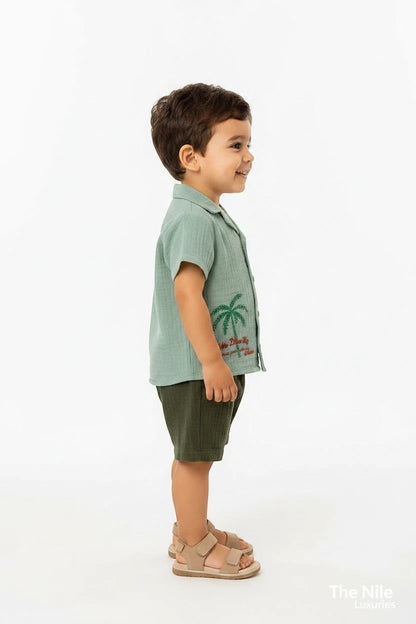 Toddler boy wearing a The Nile Luxuries sage green muslin shirt with palm embroidery and olive green shorts.