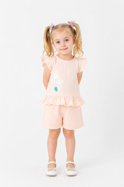 Toddler girl wearing a peach The Nile Luxuries peplum top with flutter sleeves and matching shorts with floral detail.