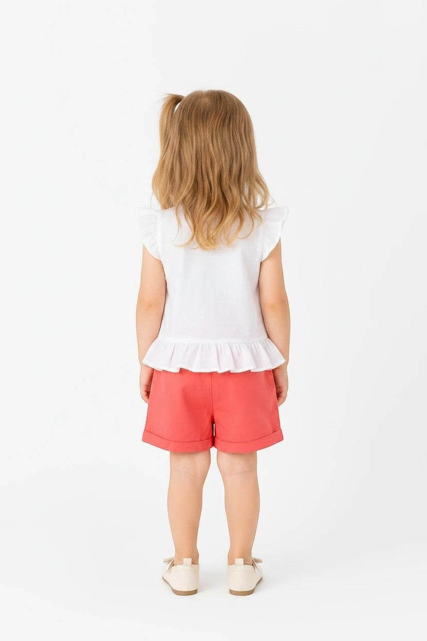Toddler girl wearing The Nile Luxuries white peplum top with flutter sleeves and coral shorts on a white background.
