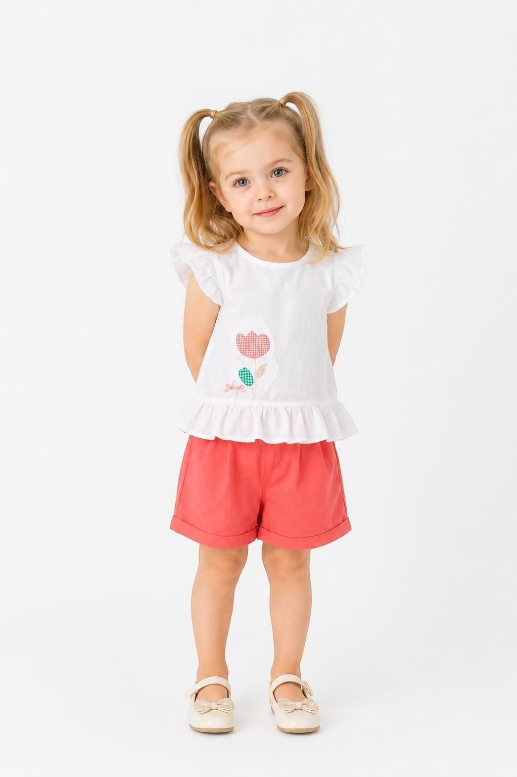 Toddler girl wearing The Nile Luxuries white peplum top with floral graphic and coral shorts on a white background.
