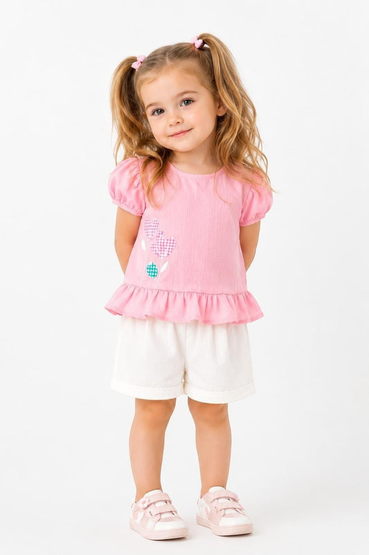 Toddler girl wearing a pink peplum top with puff sleeves and floral detail and white shorts by The Nile Luxuries