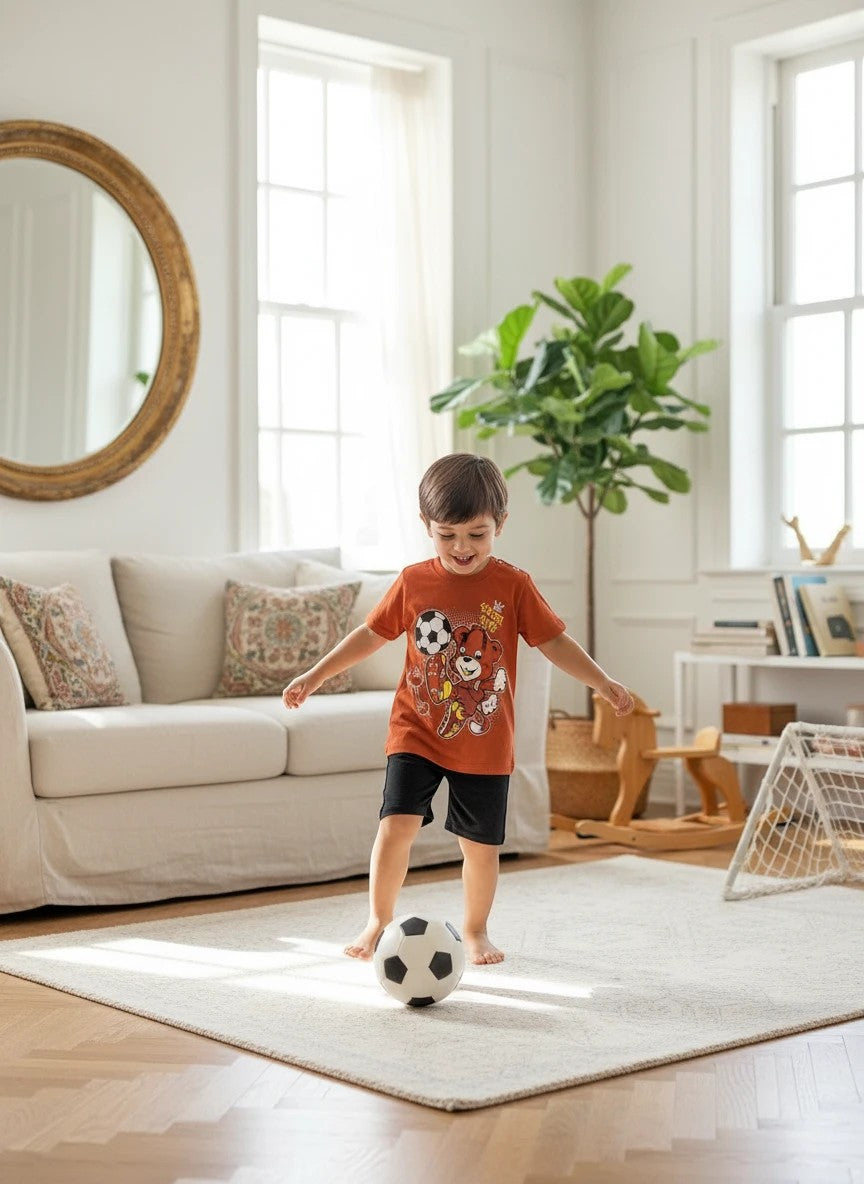 The Nile Luxuries toddler boy wearing a rust orange soccer bear graphic t-shirt and black shorts playing with a ball.