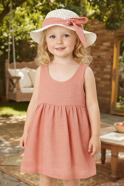 Toddler wearing a The Nile Luxuries sleeveless dusty pink dress and matching sun hat with a bow in a garden setting.
