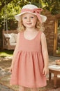 Toddler wearing a The Nile Luxuries sleeveless dusty pink dress and matching sun hat with a bow in a garden setting.