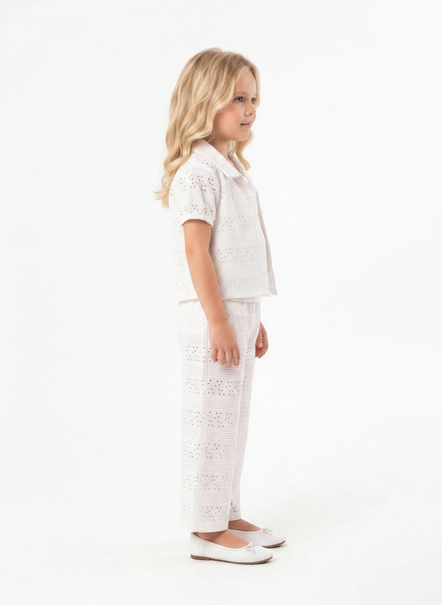 A young girl wearing a white The Nile Luxuries eyelet embroidered shirt and matching wide-leg trousers, side view.