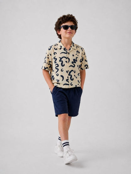 The Nile Luxuries short sleeve collared shirt with abstract navy print on cream fabric. Boy wearing shirt and navy shorts.