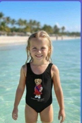 The Nile Luxuries black one-piece swimsuit for girls with a graphic teddy bear wearing a red dress and blue sash.