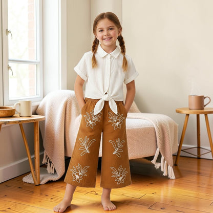 The Nile Luxuries off-white textured cropped shirt with tie front and brown wide-leg pants with white floral embroidery.