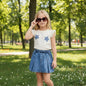The Nile Luxuries girls' cream graphic tee with floral appliques and blue denim pleated skirt set.