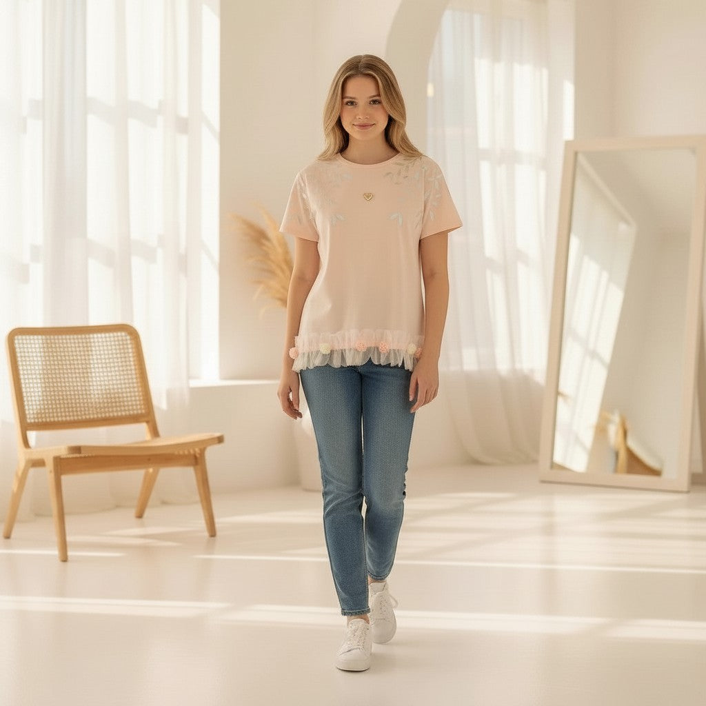 The Nile Luxuries light pink t-shirt with leaf embroidery, ruffle hem, and floral accents. Model wears jeans.